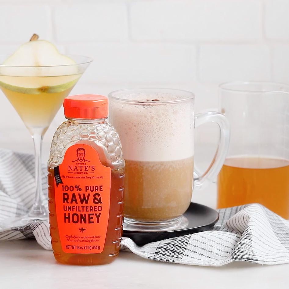 A frothy coffee latte in a mug with a bottle of Nate’s honey, a cocktail and a checkered cloth backdrop.