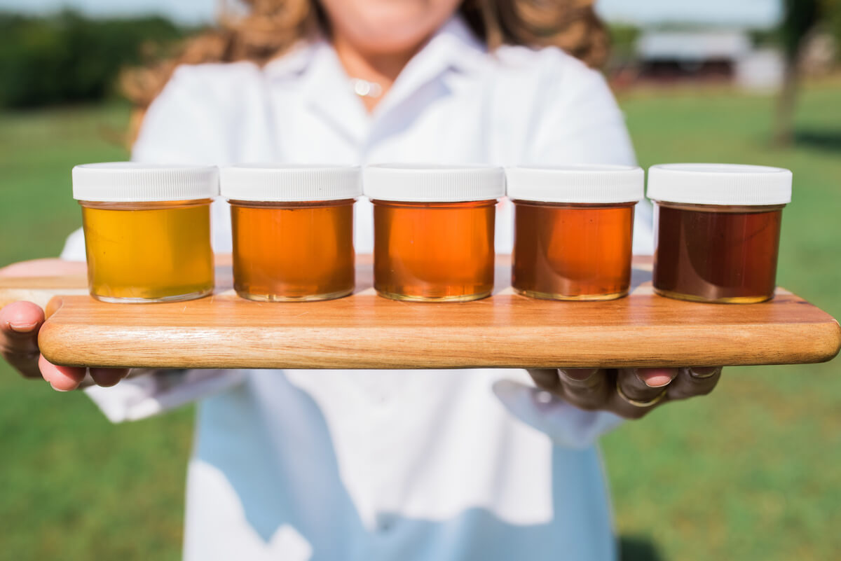 An individual in a white lab coat holding a wooden tray with five jars of honey, indicating various types and flavors.