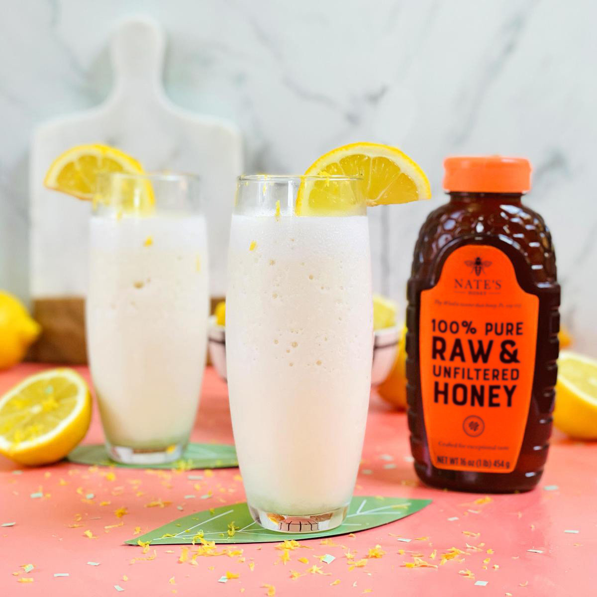 Glasses of lemonade garnished with lemon slices next to a bottle of Nate’s Honey on a speckled pink surface.