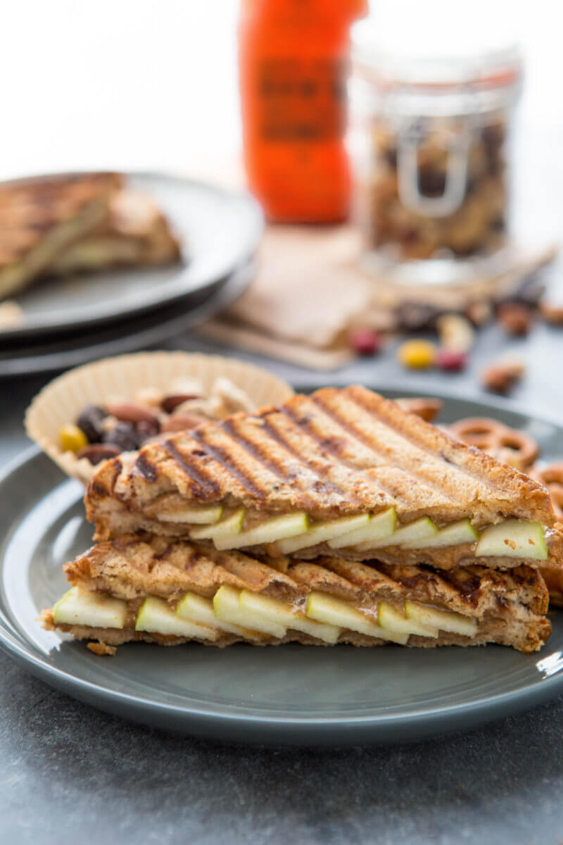 A grilled panini sandwich on a plate with apple slices and  cheese with a bottle of Nate’s honey and a jar of nuts.