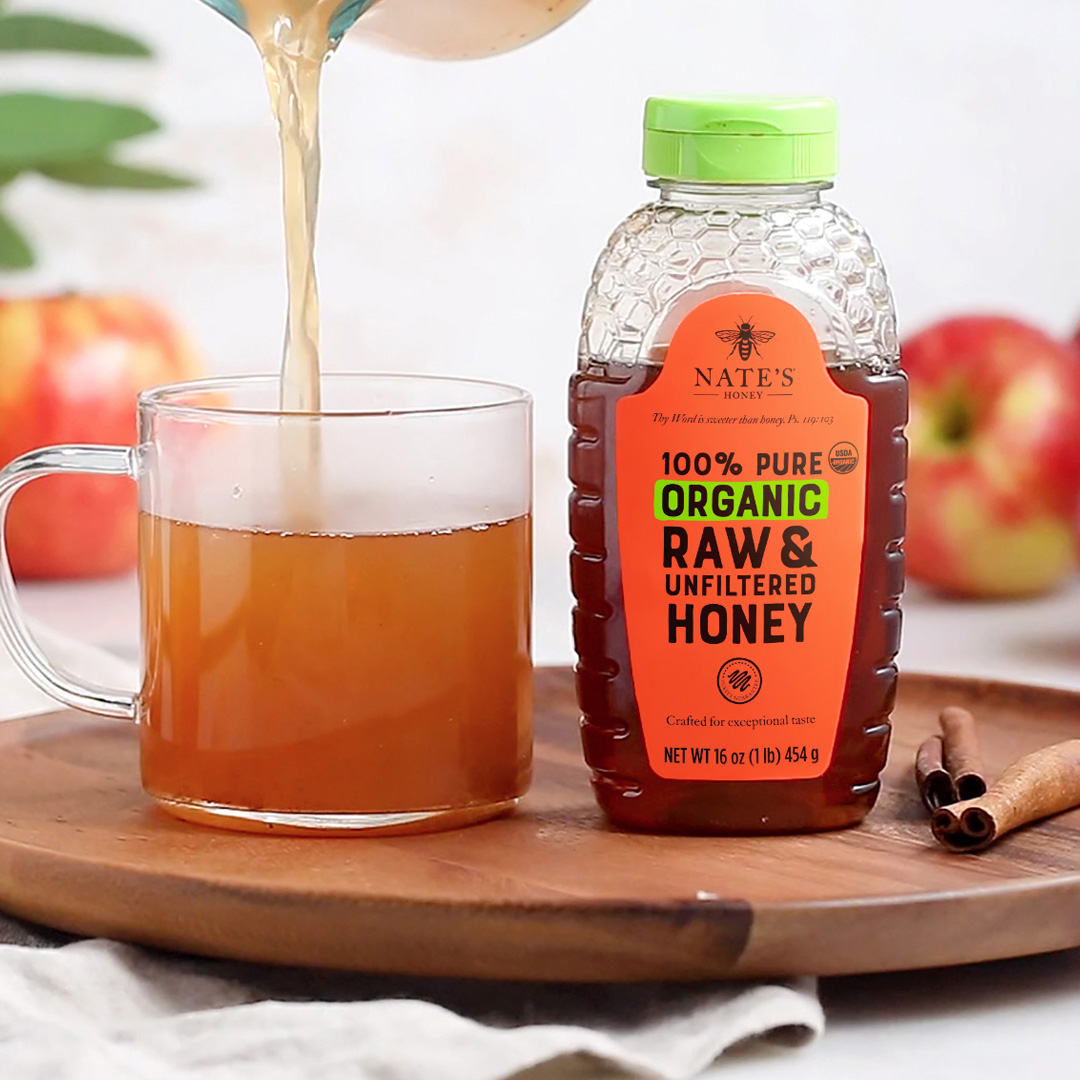 Organic Nate’s Honey bottle and apple cider being poured into a mug on a wooden tray, with cinnamon sticks and apples.