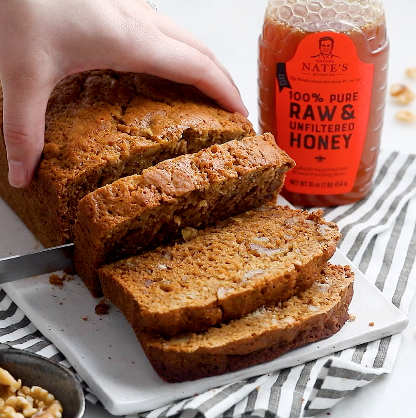 A hand slicing a loaf of bread next to a bottle of Nature Nate's 100% Pure Raw & Unfiltered Honey.