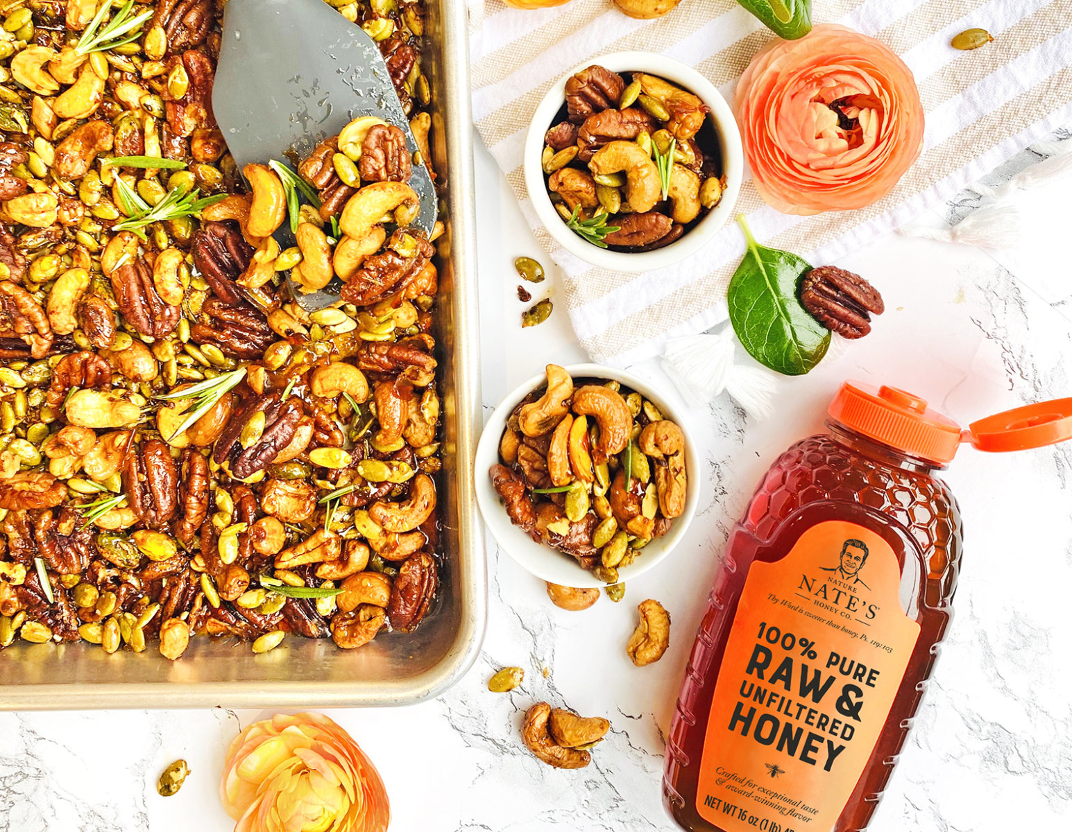 Spiced nuts mix on a tray and in bowls with a bottle of Nate's Honey, on a marble background with floral garnishes.