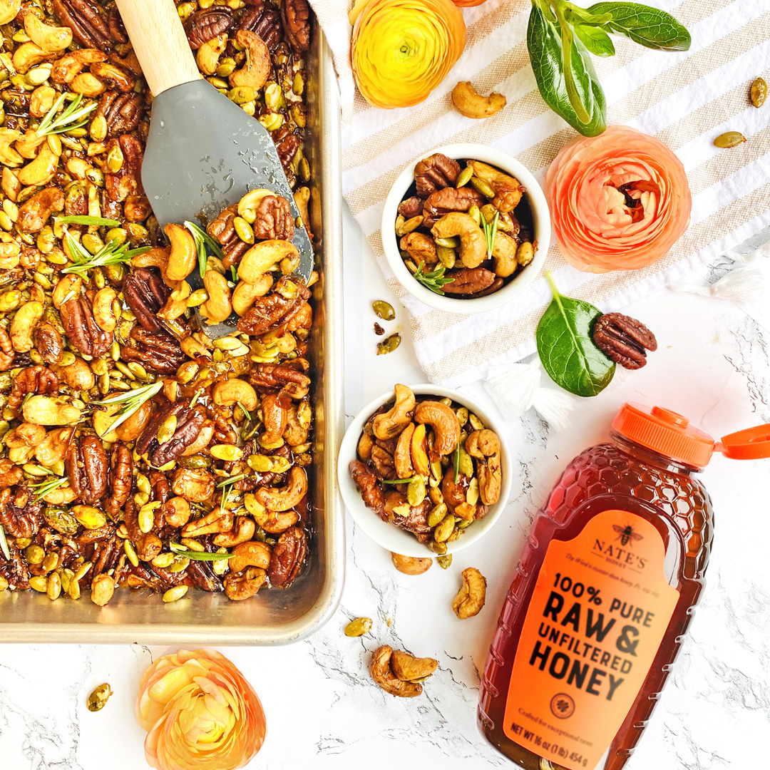 Spiced nuts mix on a tray and in bowls with a bottle of Nate's Honey, on a marble background with floral garnishes.