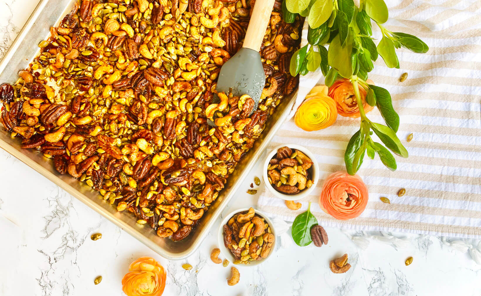 Tray of honey-roasted nuts and seeds with a spatula, beside floral decorations and greenery on a white striped cloth.