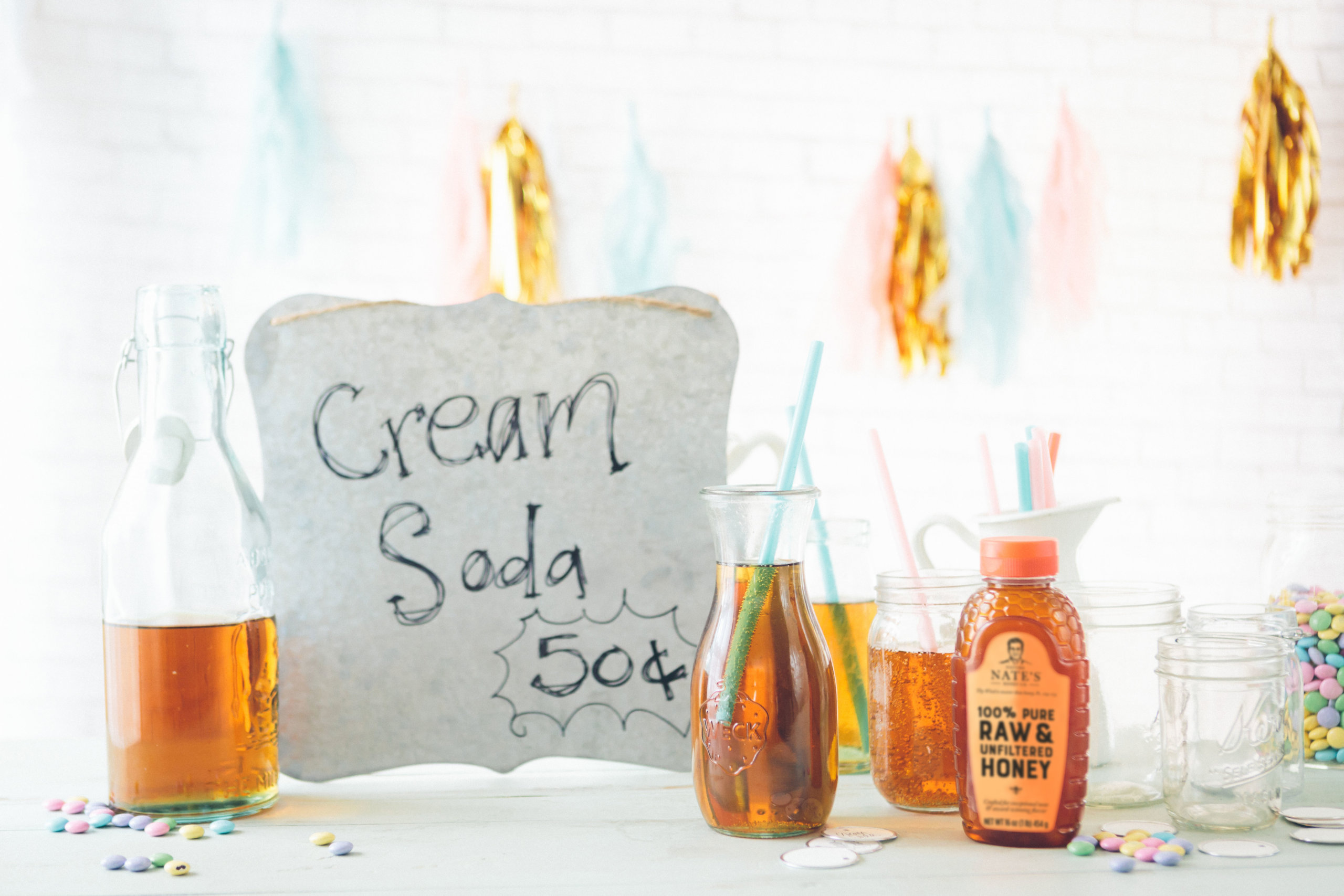 A festive setup with a Cream Soda 50¢ sign, bottles of drink, a Nate's honey bottle, straws and confetti.