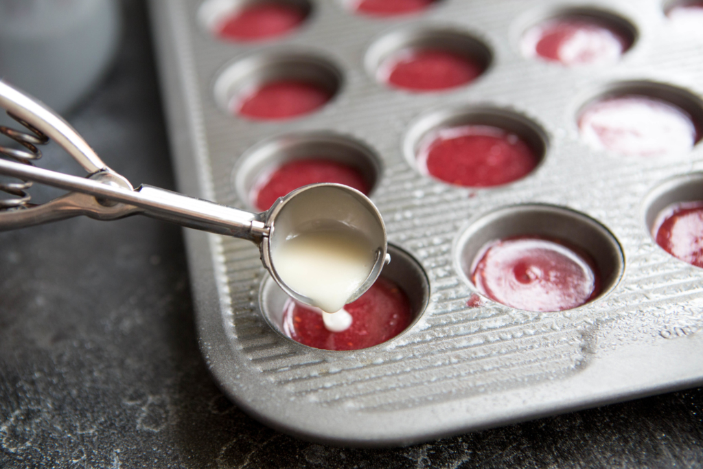 A baking pan prepped with vibrant red fruit snack mixture in each cup, with a scoop pouring creamy mix on top.