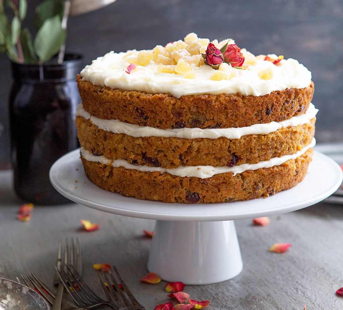 Layered carrot cake with cream cheese frosting on a cake stand, with dark floral background.