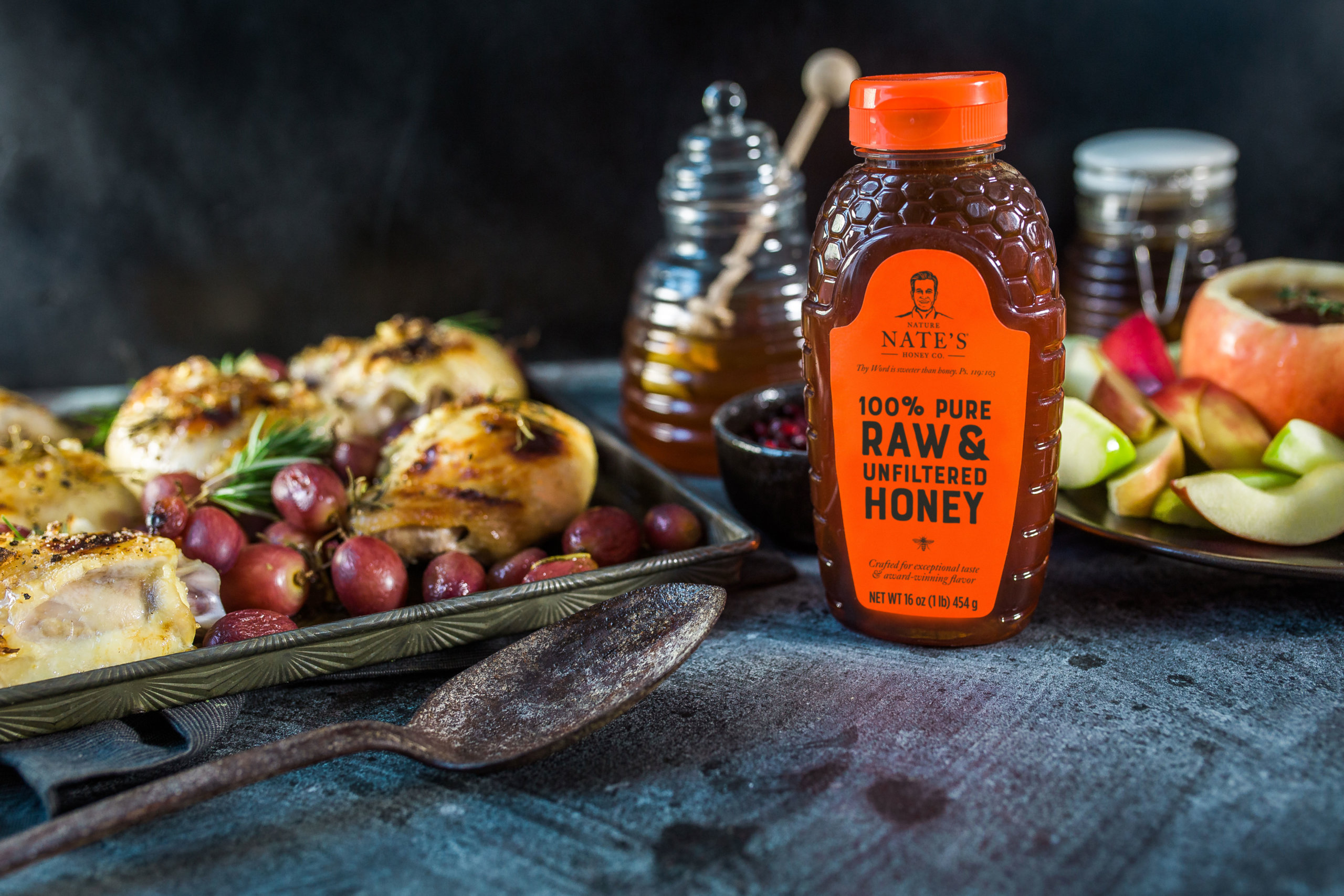 A bottle of Nate’s honey is centered on a dark surface with baked chicken, grapes and sliced apples in the background.