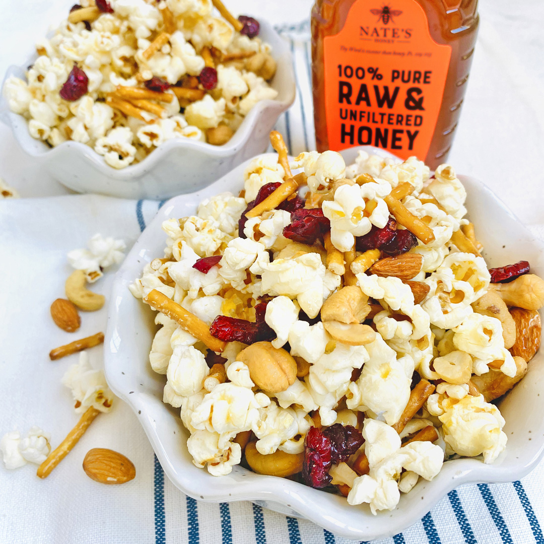 A bowl of popcorn mixed with nuts, pretzels and dried cranberries, with a bottle of Nate's Honey on the side.