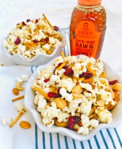 Two bowls of popcorn mixed with nuts and dried fruits, accompanied by a bottle of Nate’s raw & unfiltered honey.