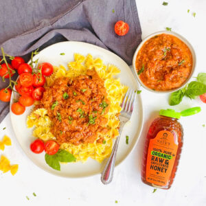 A plate of pasta with herb-garnished, Bolognese sauce, beside a Nate’s organic honey bottle, a sauce bowl and tomatoes.