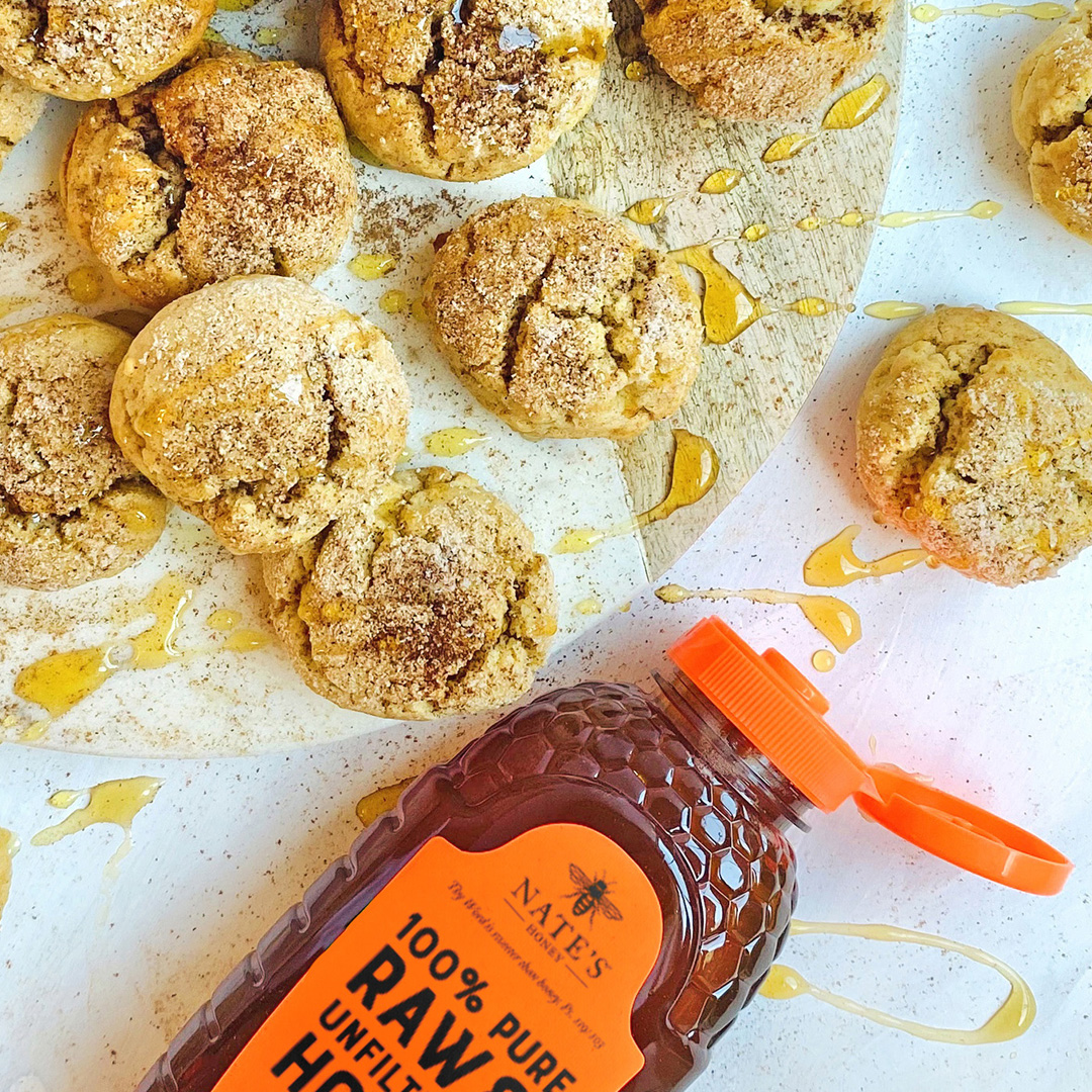 Snickerdoodle cookies drizzled with honey and a Nate's Honey bottle on a white surface with honey splatters.