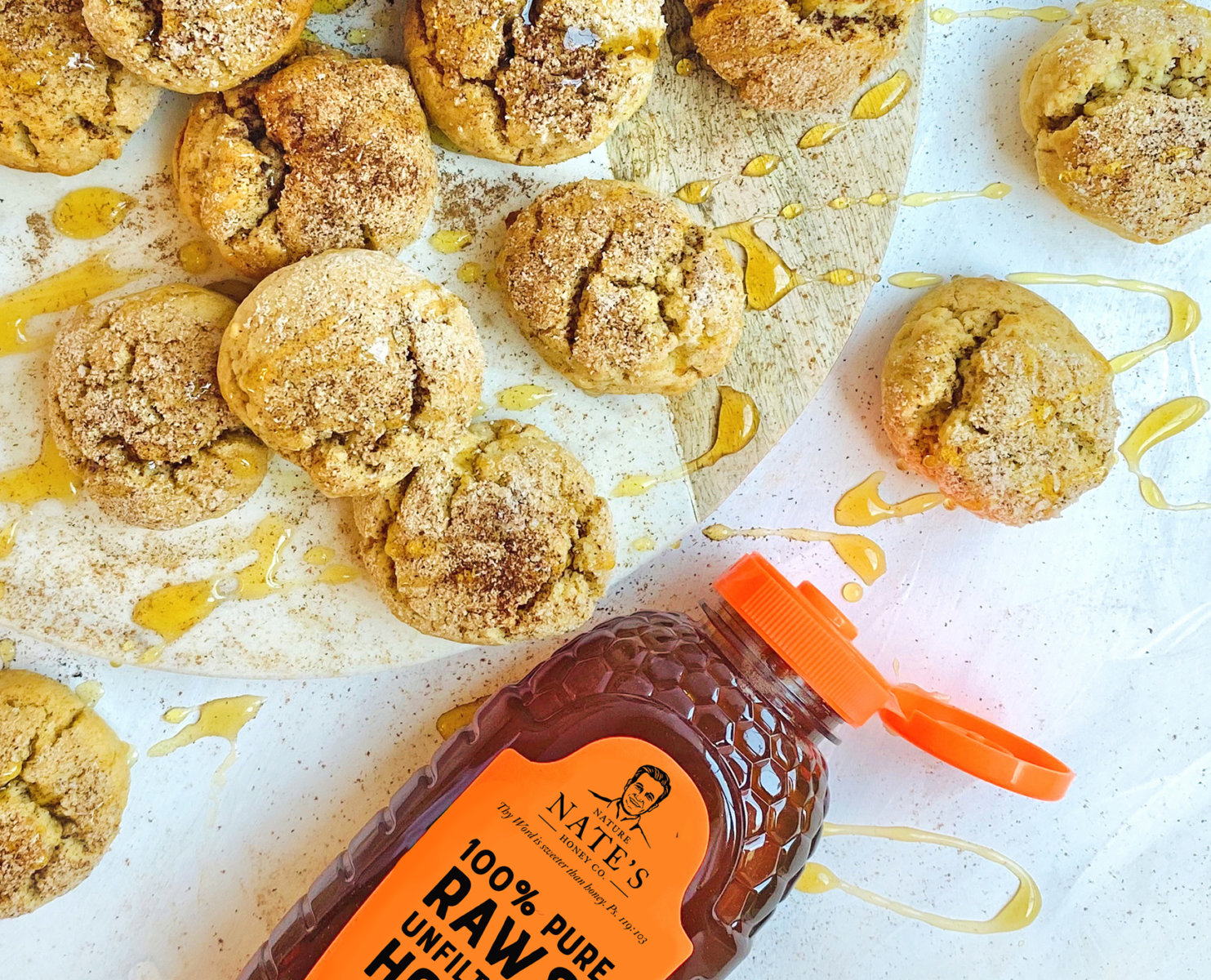 Snickerdoodle cookies drizzled with honey and a Nate's honey bottle on a white surface with honey splatters.