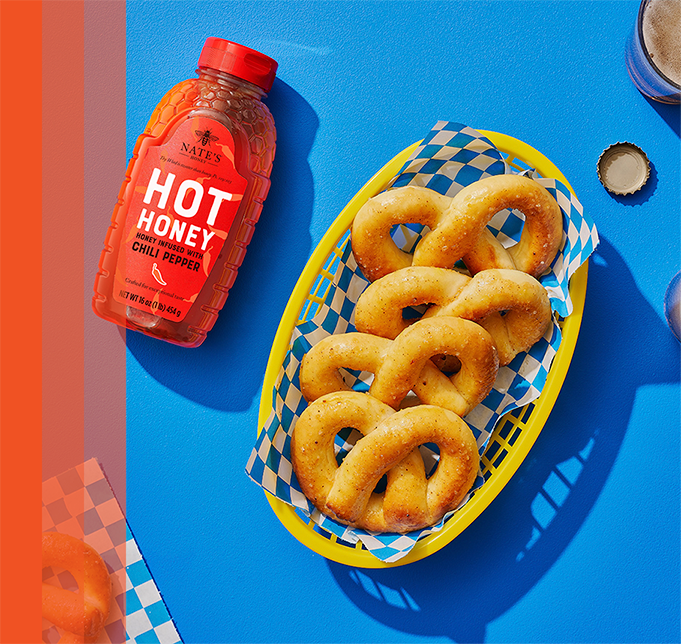 Soft pretzels in basket next to bottle of Nate's Hot Honey on blue background.