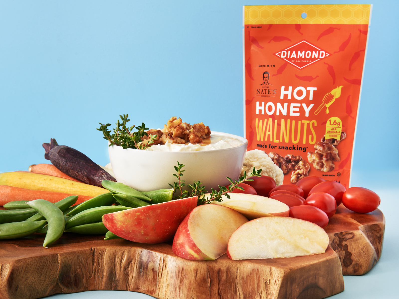 A bowl of whipped feta cheese on a board with fresh vegetables, fruits and a package of Diamond hot honey walnuts.