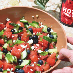 Bowl of watermelon salad with cucumber, blueberries, feta, and mint, drizzled with hot honey.