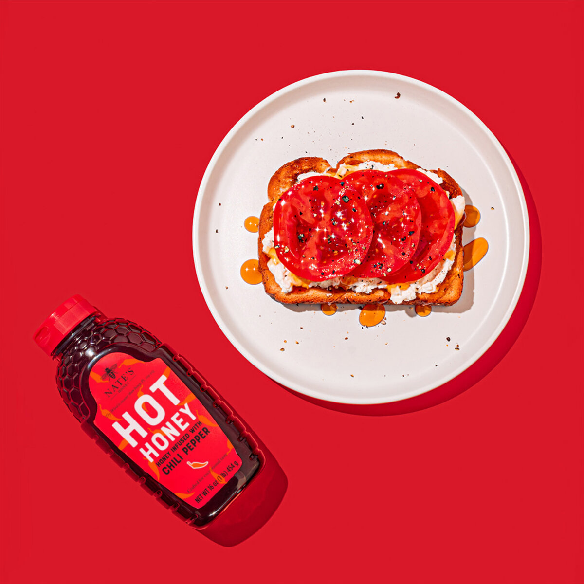 Tomato toast drizzled with hot honey on white plate with red background next to hot honey bottle.