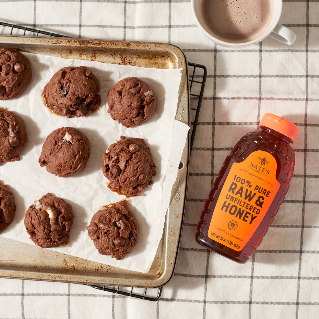 Chocolate cookies on a baking sheet with a bottle of Nate’s Honey and a cup of hot cocoa on a checked cloth.