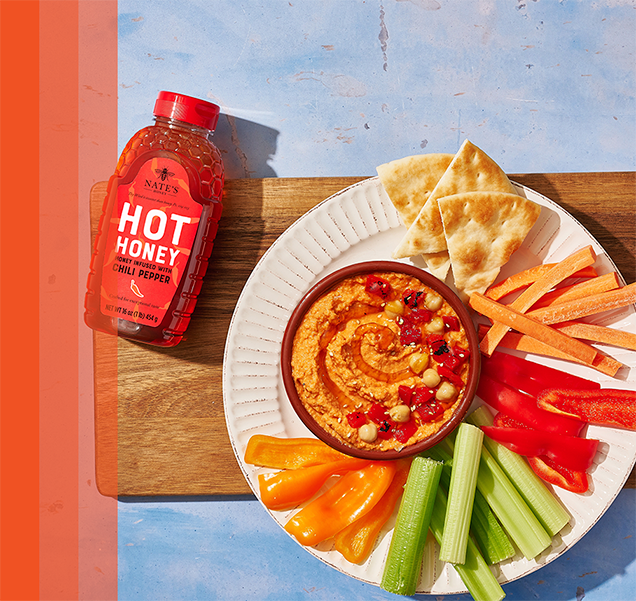 Red pepper hummus dip in a bowl surrounded by veggies and pita on a cutting board with a bottle of Nate's Hot Honey.