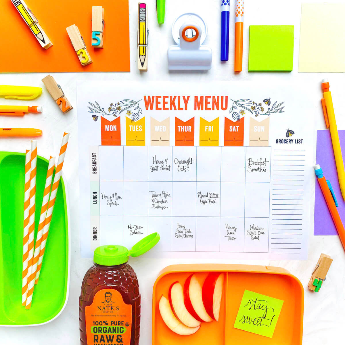 Colorful weekly menu planner with stationery, snacks, apple slices and a bottle of Nate’s honey.