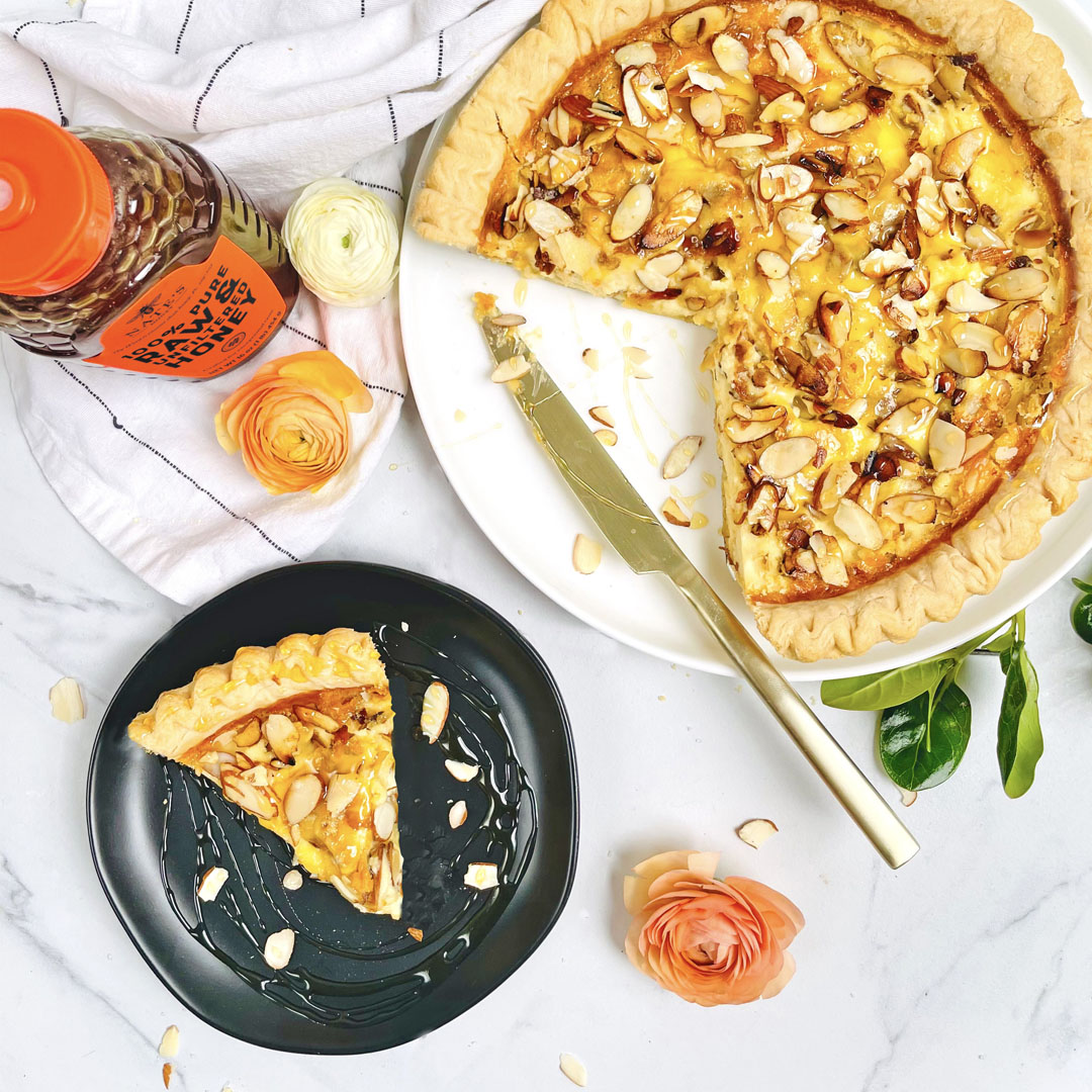 Honey Almond Sweet Quiche on platter and on black plate next to bottle of Nate's Honey, flowers and white towel on marble background