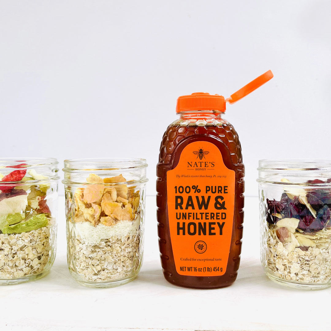Nate's Honey bottle with glass jars filled with oatmeal and assorted fruit on a white backdrop.