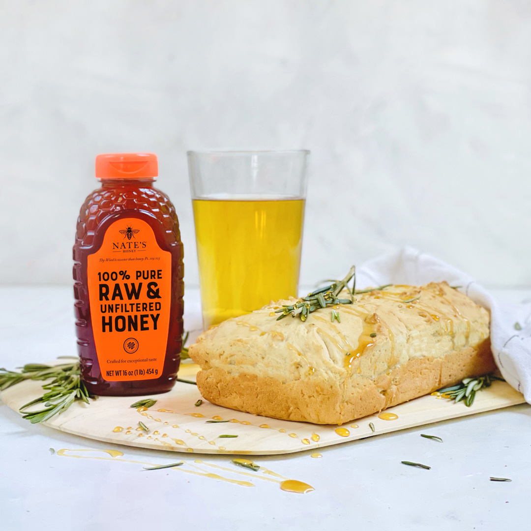 Beer bread on a board drizzled with honey and garnished with rosemary sprigs, a Nate’s Honey bottle and a glass of beer.