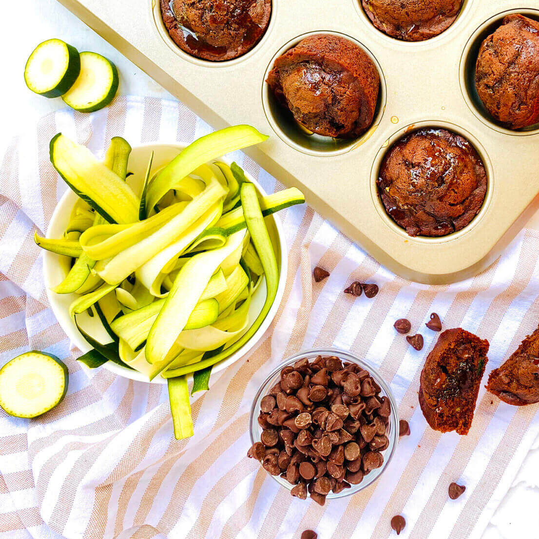Chocolate muffins in a pan, Nate’s honey bottle, chocolate chips and zucchini ribbons on a striped towel.