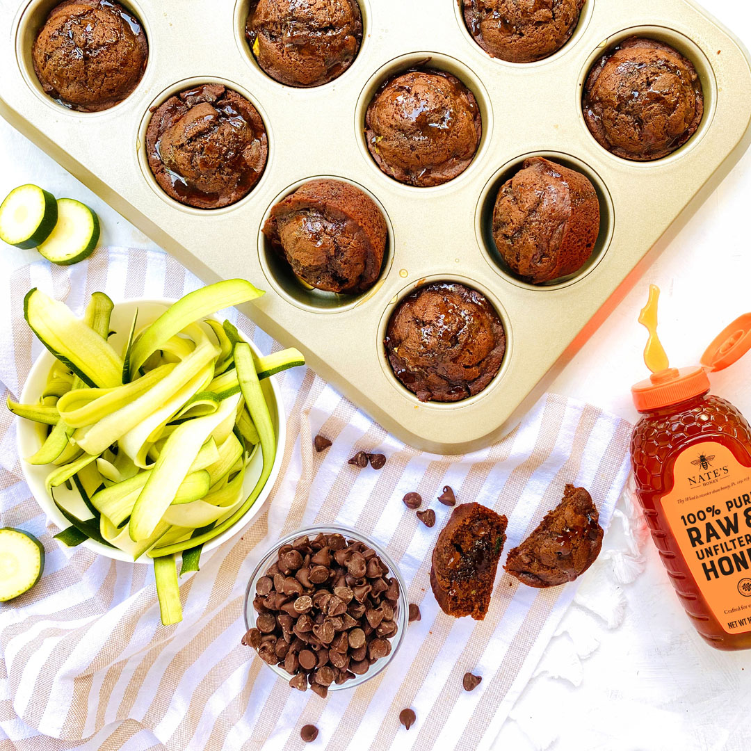 Chocolate muffins in a pan, Nate’s Honey bottle, chocolate chips and zucchini ribbons on a striped towel.