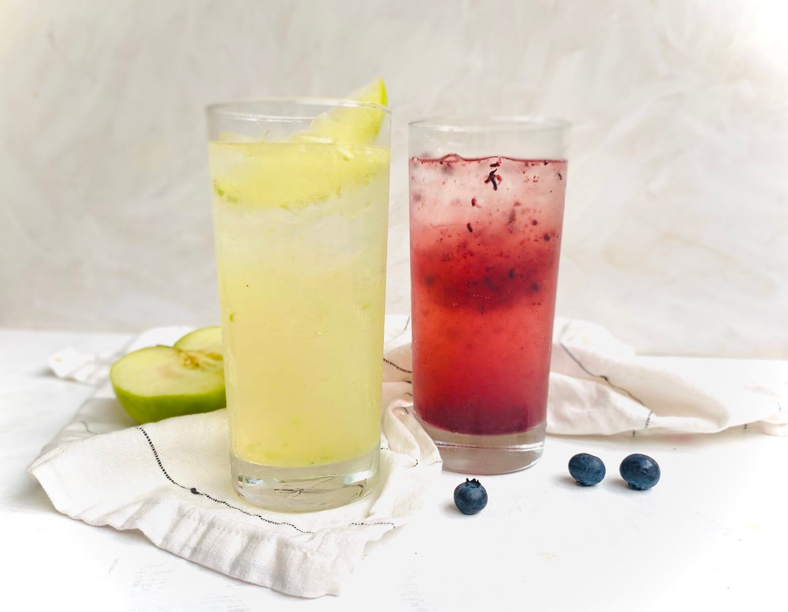 Two chilled drinks in tall glasses, yellow and red, with fruit garnish, beside blueberries and apple on a draped cloth.