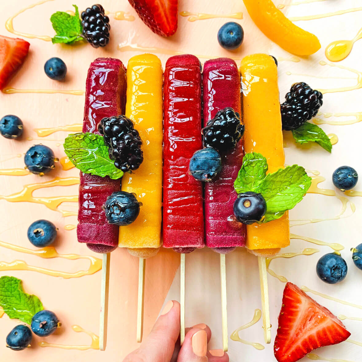 Hand holding fruit popsicles with fresh berries and mint on a drizzled honey background.