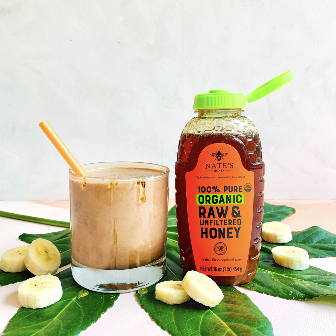 Chocolate and banana smoothie in a glass, Nate's Honey organic bottle with banana slices on a leafy backdrop.
