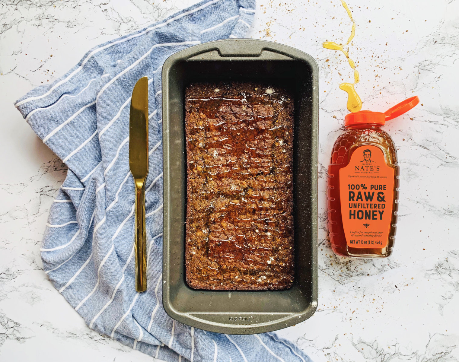 Loaf of honey whole wheat bread in a pan, knife, Nate's honey bottle and striped napkin on a marbled counter.