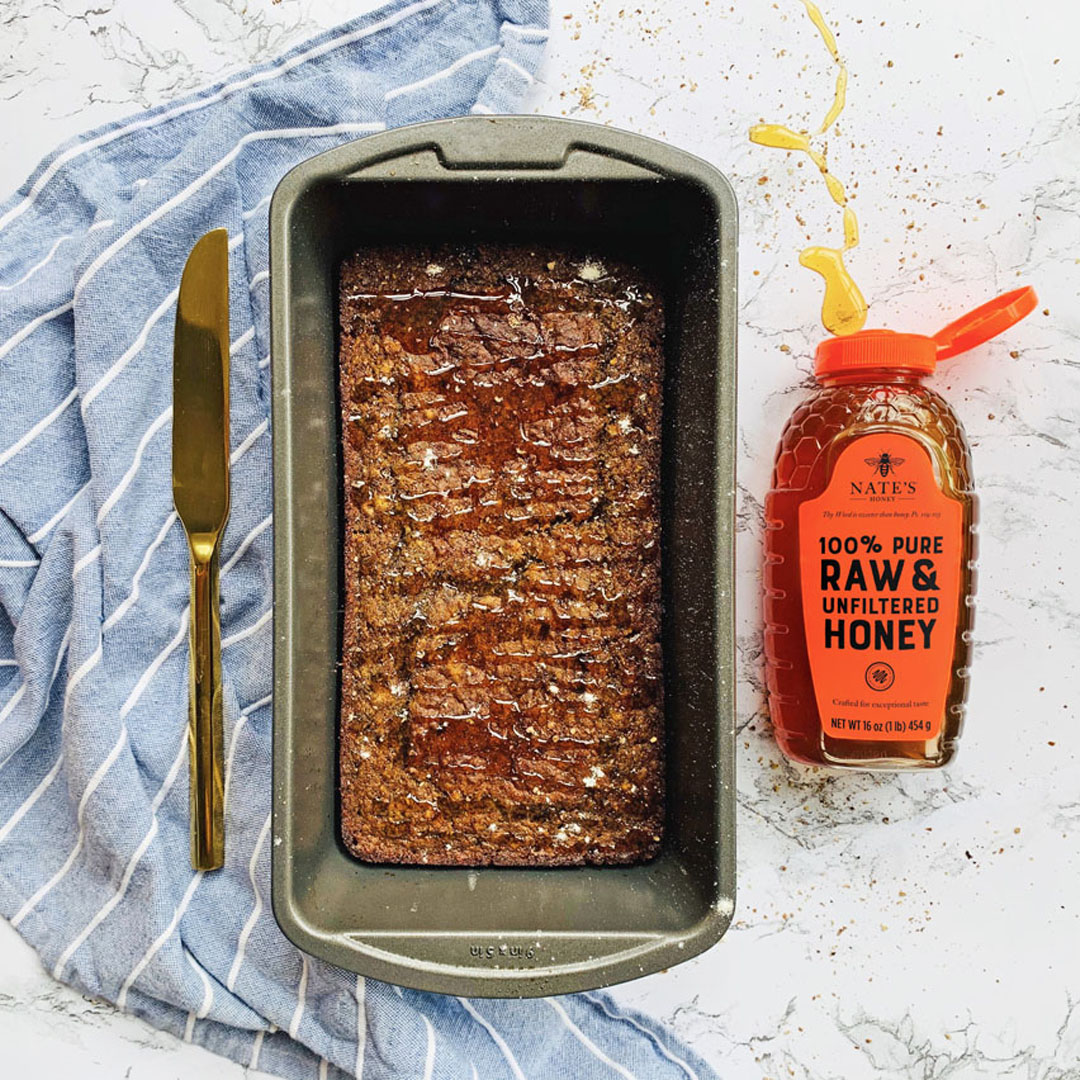 Loaf of honey whole wheat bread in a pan, knife, Nate's Honey bottle and striped napkin on a marbled counter.