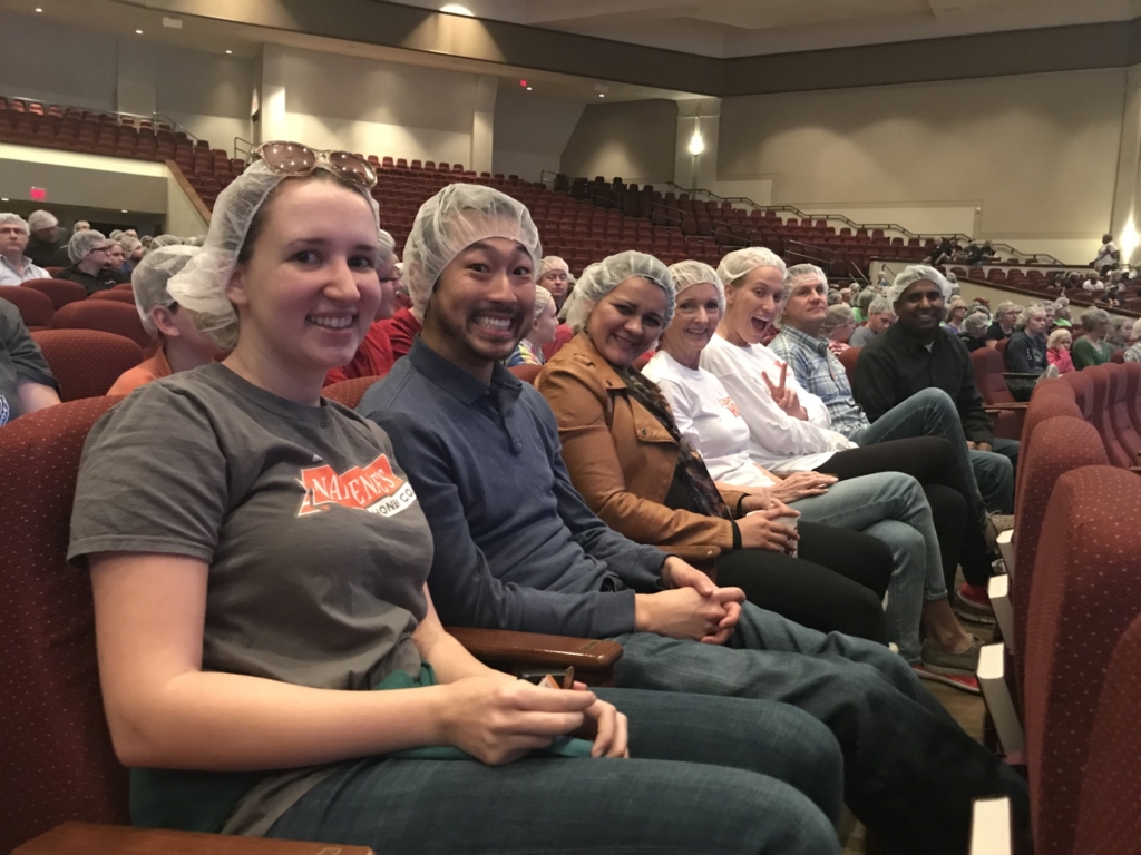 A group of people, all wearing hairnets, is sitting in rows of an auditorium, smiling and facing the camera.
