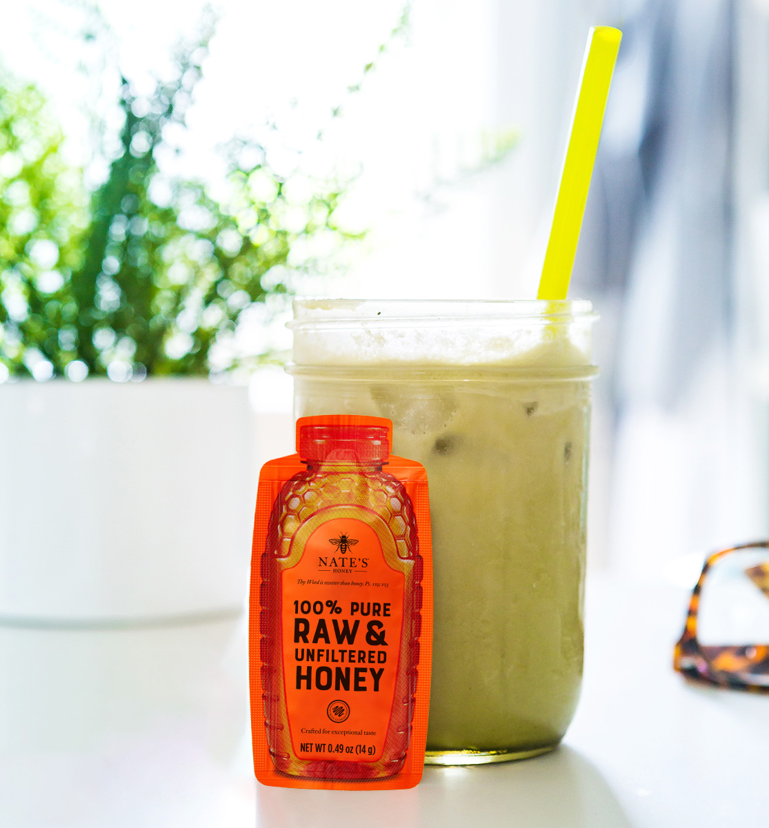 A green iced honey matcha with a yellow straw, alongside a Nate's  Honey Mini and sunglasses, with airy background.