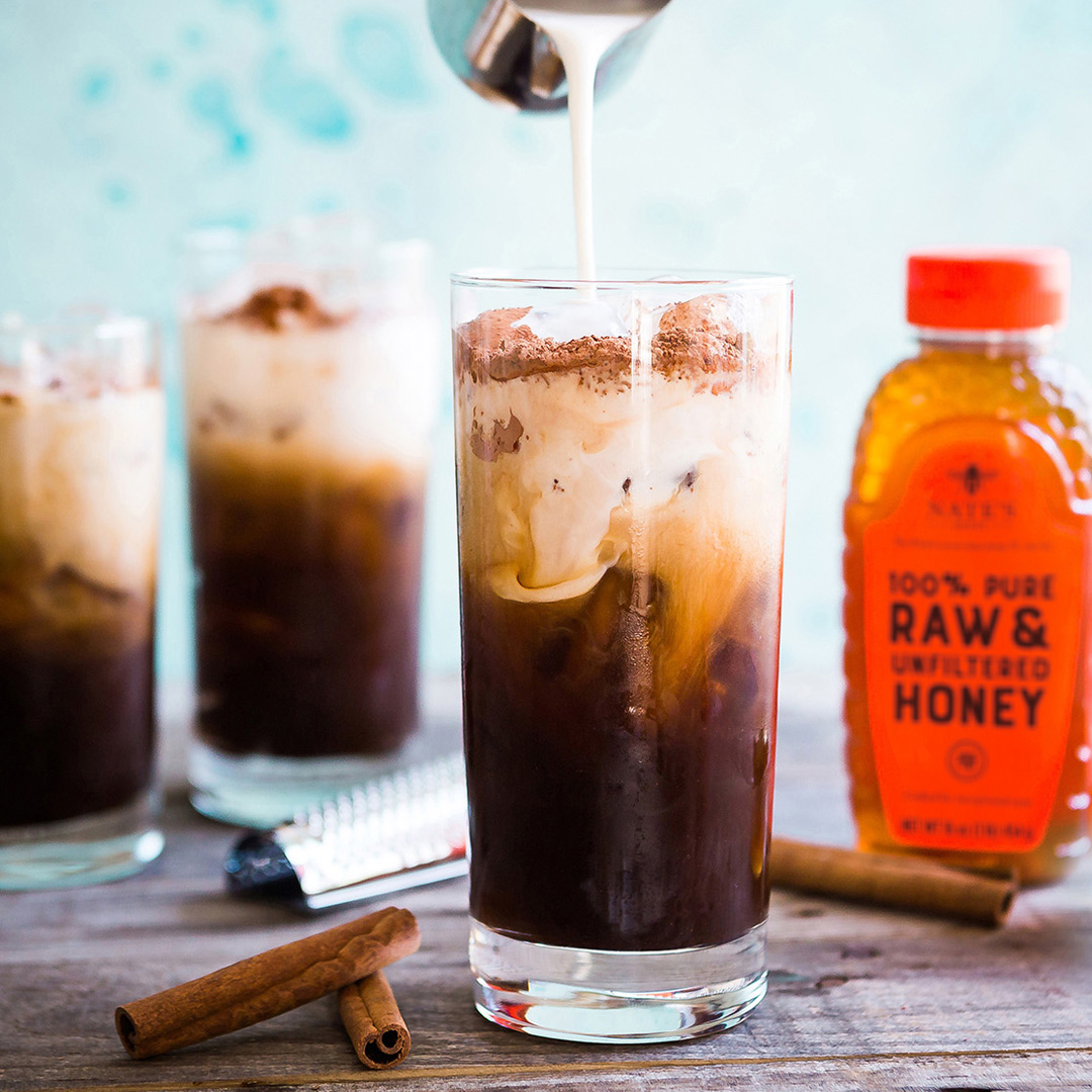 Pouring cream into iced coffee, with more glasses, cinnamon and a bottle of Nate's Honey.