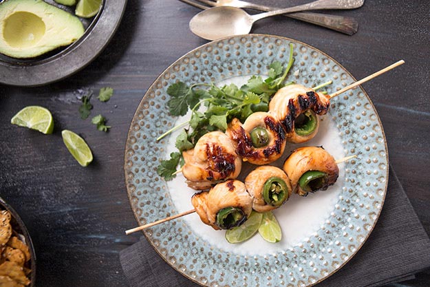Grilled chicken and jalapeño skewers on a speckled blue plate, dark wood table with lime wedges.
