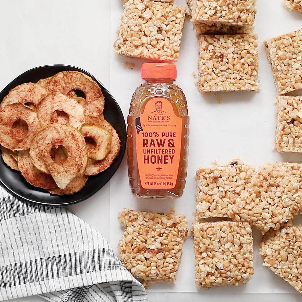 Apple chips in a black bowl, rice crispy treats, and a bottle of Nate's honey on a striped cloth.