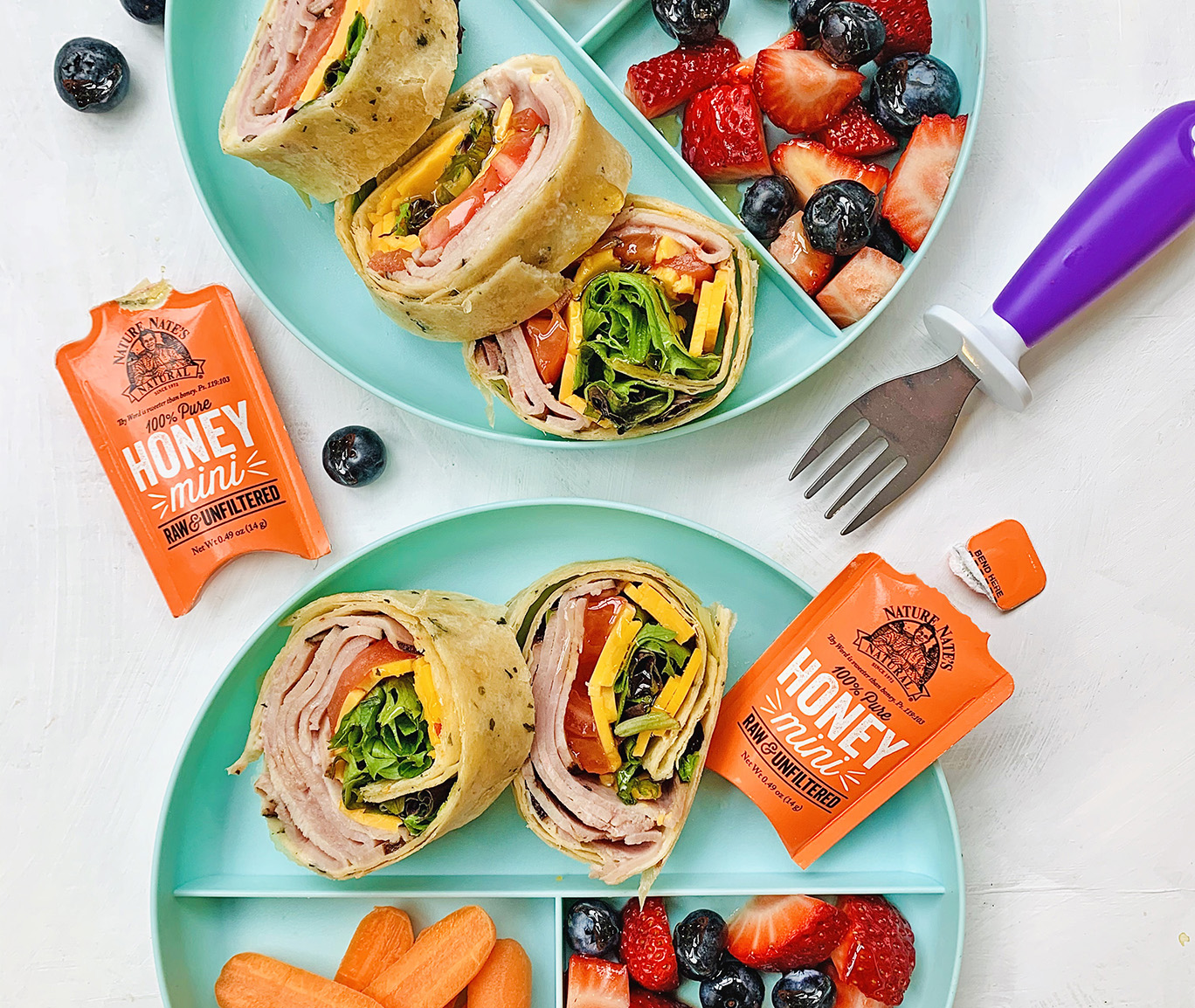 Turkey wraps on a plate with Nate's honey minis, fruit salad and carrots on the side.