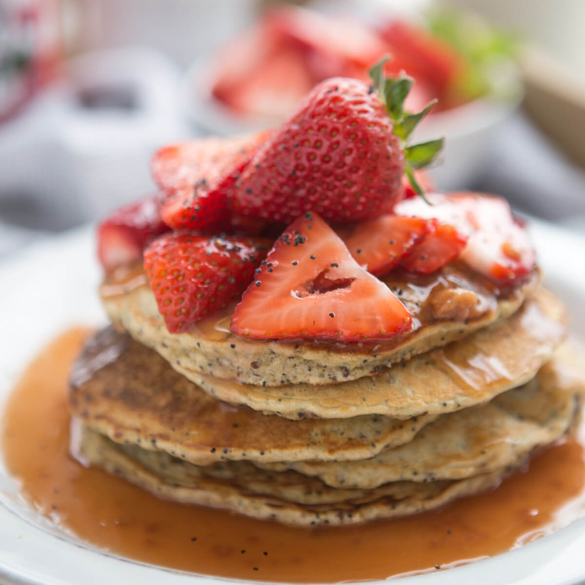 A stack of poppyseed quinoa pancakes topped with fresh, sliced strawberries and drizzled with golden honey.