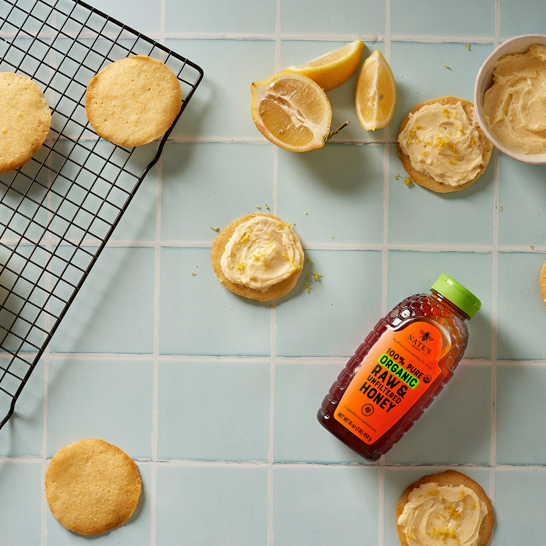 Lemon frosted cookies, fresh lemon slices, and a bottle of organic Nate’s Honey on a blue tiled countertop.