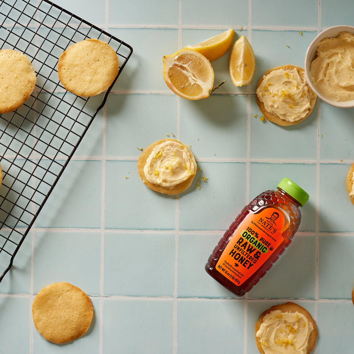 Lemon frosted cookies, fresh lemon slices, and a bottle of Nate’s organic honey on a blue tiled countertop.