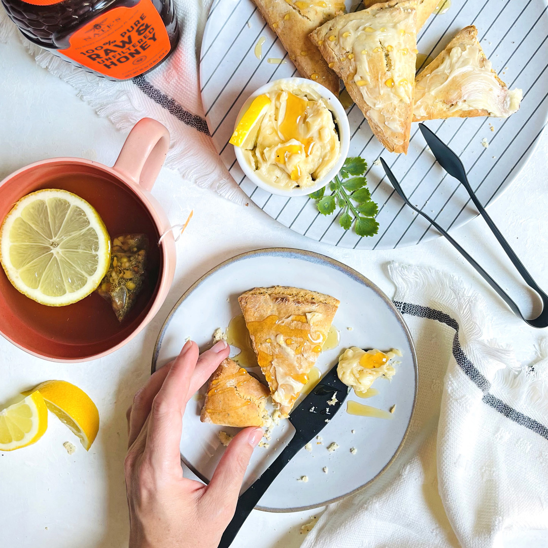 Lemon poppyseed scones drizzled with honey on a plate, a cup of tea, and a bottle of Nate’s Honey on a white surface.