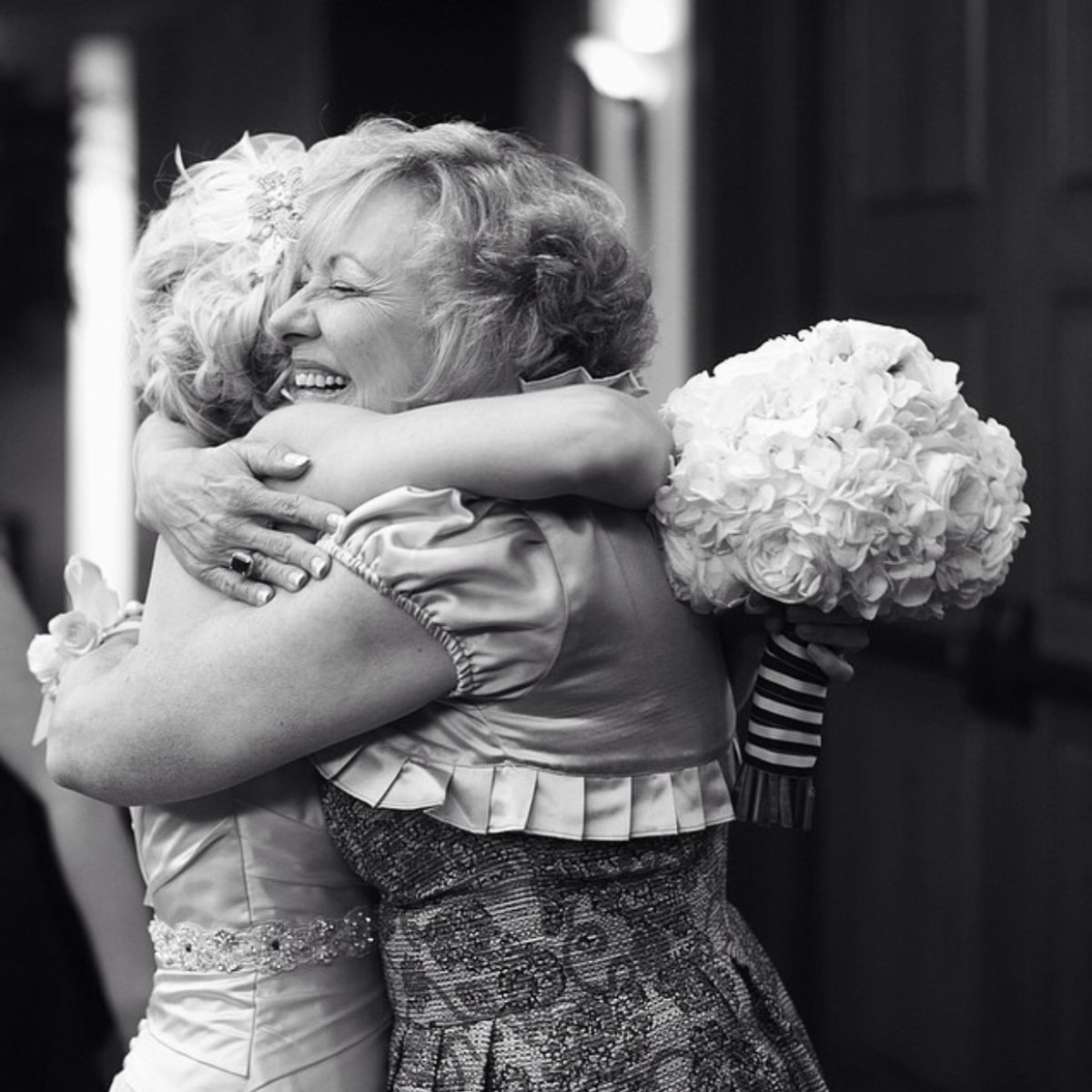 A black and white photo of two women embracing in a heartfelt hug, one holding a large bouquet of flowers.