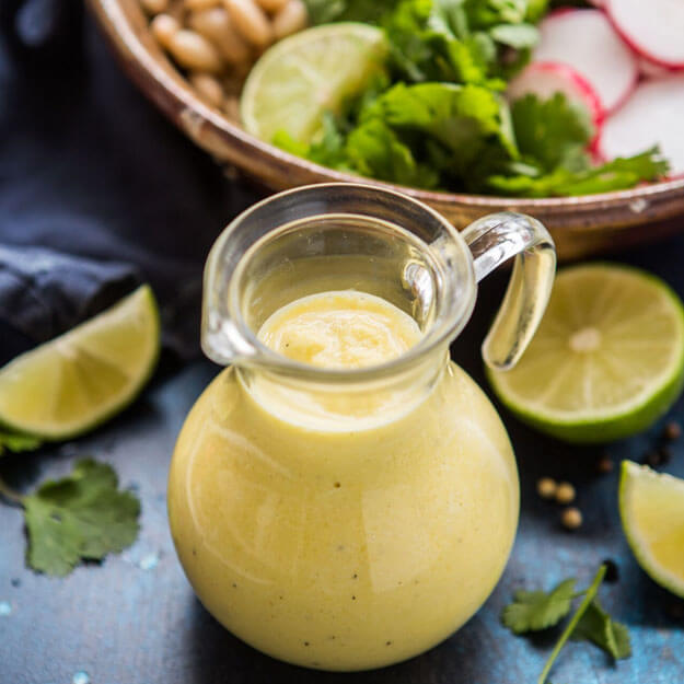 Creamy mango and lime salad dressing in a small pitcher, halved limes, cilantro around, dark moody setting.