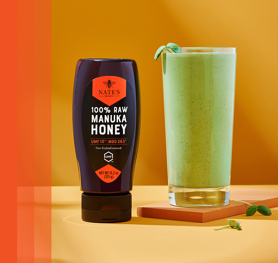 Nate's New Zealand Manuka Honey bottle beside a green smoothie on a yellow surface with an orange background.