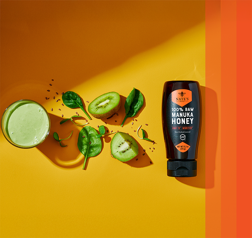 Manuka honey bottle next to a green smoothie on a yellow background.