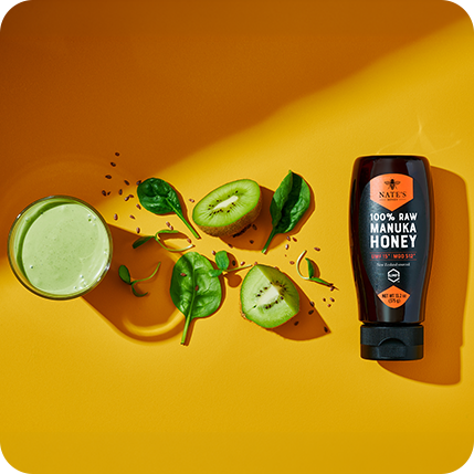 Nate’s Manuka Honey bottle with kiwi slices, spinach, and a green smoothie on a bright yellow background.